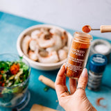 A hand holds a small glass jar of smoked paprika seasoning above a turquoise countertop, with a spoonful of the orange-red spice hovering above the jar. A bowl of shrimp and other cooking ingredients are blurred in the background.