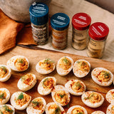 Four seasoning bottles from Morton's & Bassett Company arranged on a wooden surface behind deviled eggs garnished with dill and red pepper flakes. The bottles feature blue and red caps and are labeled with flavors including Cumin, Smoked Pepperoni, Smoked Paprika, Onion Powder, and Garlic Powder.