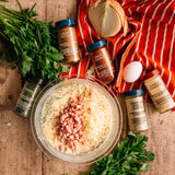 A styled flat-lay of a glass bowl filled with a creamy yellow mixture topped with chopped pink meat and shredded cheese, surrounded by seasoning bottles, a red-and-white striped cloth, parsley, an onion, garlic powder, and an egg on a wooden surface.