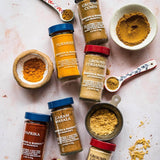 An overhead flat-lay of Morton & Bassett ground spices, including Turmeric, Ground Cumin, Ground Coriander, Paprika, Caram Masala, and Ground Ginger, arranged with measuring spoons and bowls on a light pink marble background.