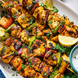 Grilled chicken and vegetable skewers on a white serving platter, garnished with chopped cilantro and lemon slices, with a small bowl of white dipping sauce in the corner.
