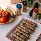 Ground cumin and coriander spice blends in glass jars with blue, black, and red lids, arranged on a kitchen counter with cooked skewered meat, tomatoes, cucumbers, and lemon nearby.