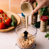 A hand pours a creamy beige dressing from a small beige pitcher into a clear food processor bowl filled with chickpeas, with tomatoes, a cucumber, and spice jars in the background.