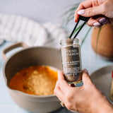 A person holds a small glass jar of ground cardamom with a black scoop over a pot of ingredients, showing the spice blend being used in cooking.