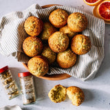 A top-down styled food photo showing a basket of golden-brown muffins lined with a striped cloth, with two spice jars and sliced citrus fruit on a light countertop. One muffin is broken in front to show the crumb texture.