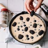 A cast iron skillet filled with a light beige batter topped with fresh blackberries and blueberries, set on a striped cloth over a white tiled surface. A hand is placing a blackberry into the skillet, with a spice jar and bottle visible in the background.