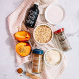 An overhead flat-lay of a beverage mix or recipe ingredients arranged on a pale pink striped cloth over a white surface. Visible items include a bottle of vanilla extract, bowls of oats and shredded powder, a jar of ground cinnamon, a jar of ground almonds, and sliced peaches.