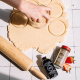 A hand uses a glass cookie cutter over rolled-out dough on a white tiled surface, with a wooden rolling pin, vanilla extract bottle, spice jar, and strawberries nearby.