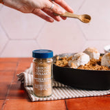 A glass jar of Morton & Bassett Ground Ceylon Cinnamon spice sits on a striped cloth on a terracotta tile surface, with a hand holding a wooden spoon sprinkling spice above a skillet containing baked goods.