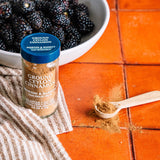 A bottle of Morton & Bassett Ground Ceylon Cinnamon spice jar with a blue lid, placed on a striped cloth on an orange tiled surface, with a wooden spoon of cinnamon and a bowl of blackberries in the background.