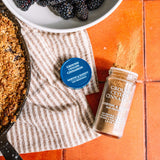 A jar of Morton & Nasset Ground Ceylon Cinnamon next to its blue screw cap lid, styled on a terracotta tiled surface with a striped cloth, a skillet of food, and a bowl of blackberries.
