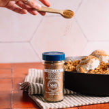 A jar of MCB Organic Ground Ceylon Cinnamon sits on a striped kitchen towel next to a baking dish with baked goods, while a hand holds a wooden spoon sprinkling cinnamon above.