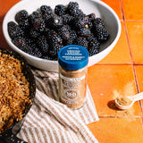 A bottle of Morton & Bashett Organic Ground Cinnamon placed on a striped cloth beside a bowl of blackberries and a skillet with baked granola on an orange tiled surface.