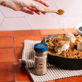 A jar of M&B Organic Ground Ceylon Cinnamon sits on a striped kitchen towel on a terracotta tile countertop, with a hand sprinkling cinnamon from a wooden spoon over a baked dish in the background.
